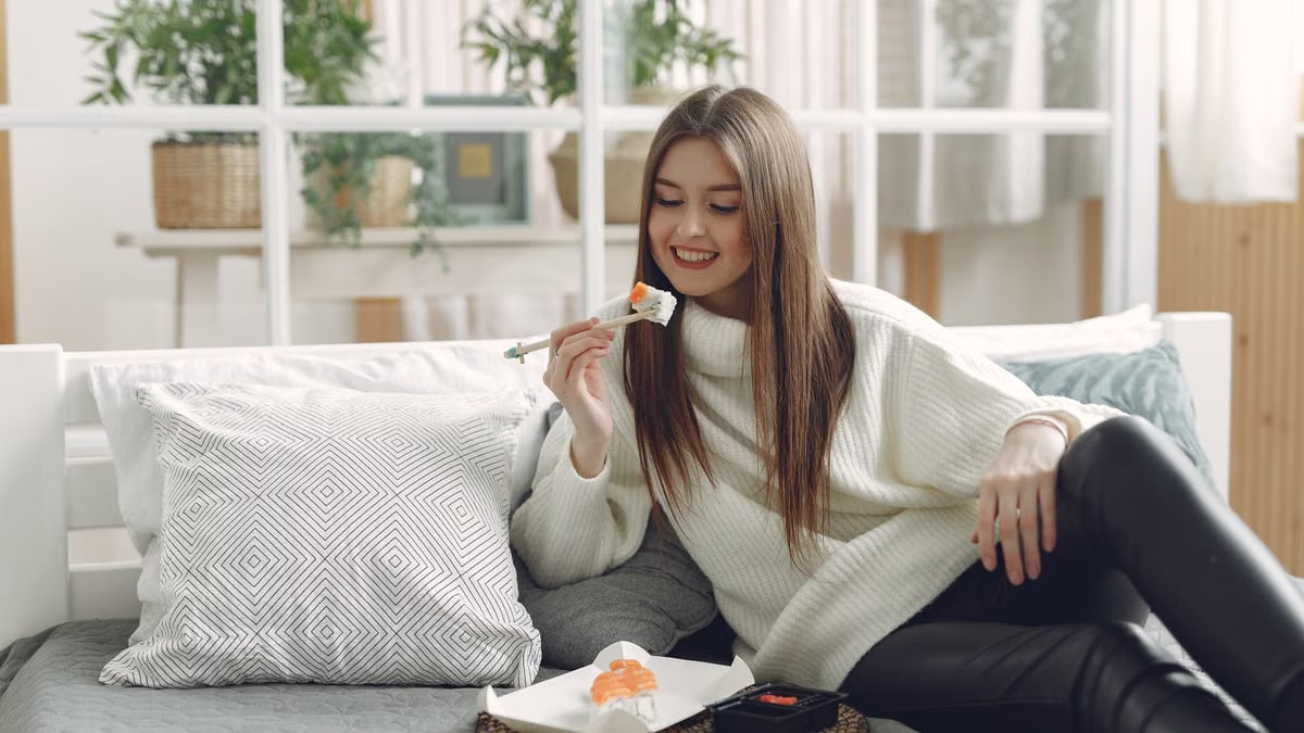 Smiling young lady in warm knitted sweater enjoying fresh sushi on wicker tray while relaxing on cozy sofa with soft cushions in modern living room